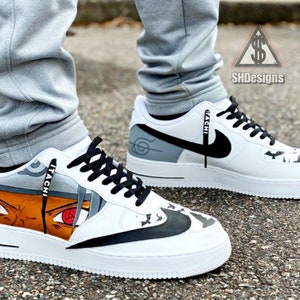 May include: White custom sneakers with a black swoosh and a black and white graphic of a character from the anime series Naruto. The character's name "ITACHI" is written on the side of the shoe.