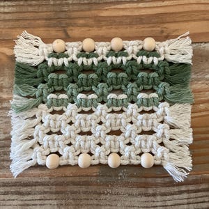 May include: A rectangular macrame wall hanging in shades of white and green, with wooden beads. The handmade piece features a geometric pattern with knotted details and fringe edges. The color palette includes light and dark green, and off-white.