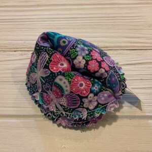 May include: A fabric fortune cookie with a floral pattern in shades of pink, purple, and green on a black background. The cookie is folded in half and has a white seam around the edge.