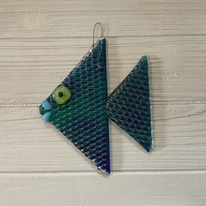 May include: A fused glass fish ornament featuring a textured, scale-like pattern in shades of deep blue and teal. The fish has a bright lime green eye with a black center and two small, light blue glass beads representing the mouth and lips.