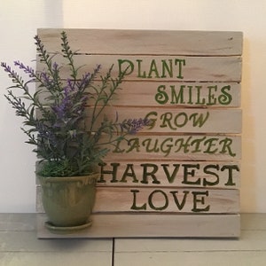 May include: A wooden sign with the words "Plant Smiles Grow Laughter Harvest Love" painted in green on a white background. A small potted plant with purple flowers is in front of the sign.