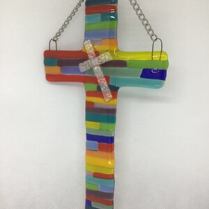 May include: A colorful glass cross with a rainbow of colors, including red, orange, yellow, green, blue, purple, and white. The cross is made of small pieces of glass that are fused together. It is hanging from a silver chain.