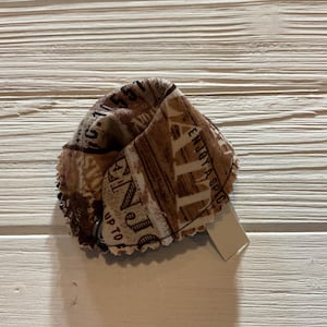 May include: A brown and white fabric heart-shaped ornament with a scalloped edge. The fabric has a vintage newspaper print with the text "Enjoy a Special Day" and "Up to 10".