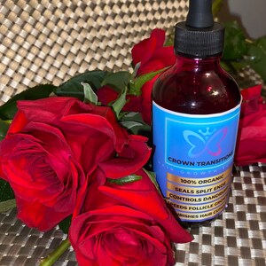 May include: A bottle of Crown Transitions hair growth elixir with a blue label and a butterfly logo, next to a bouquet of vibrant red roses. The label highlights the product's organic nature and benefits like sealing split ends and controlling dandruff.