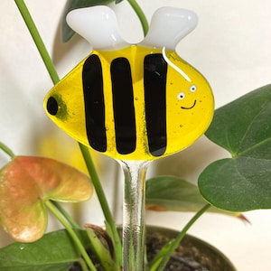 May include: A glass bee-shaped plant stake with a yellow body, black stripes, white wings, and a smiling face. The stake is inserted into a potted plant.
