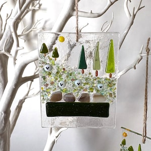 May include: A clear, rectangular glass ornament with a landscape design. The ornament features green trees, a yellow sun, and a green and brown landscape. It is suspended by a natural twine cord. The background is a white tree.