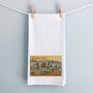 May include: A white kitchen towel with a vintage postcard design featuring the text "Greetings from Missouri" with a colorful illustration of the state's skyline. The postcard also includes the text "Seein's Believin'" and "Blue  Vine."