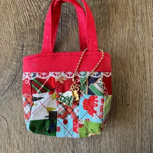 May include: A small, red tote bag with patchwork fabric in Christmas colors. The bag has a white lace trim and a gold charm with a Christmas tree. The bag has red handles.
