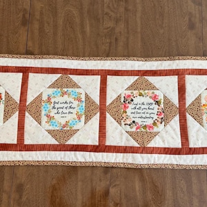 May include: A rectangular table runner with four square panels. Each panel features a floral wreath design and a Bible verse. The runner has a cream background with brown and tan accents and a brown border.