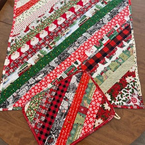 May include: Christmas-themed quilted table runner and matching pot holder. The runner and pot holder feature diagonal stripes of festive fabrics with Santa Claus, snowflakes, and plaid patterns in red, green, and white. The pot holder has a loop for hanging.