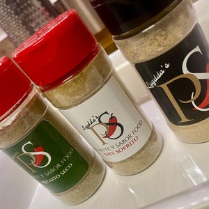 May include: Three spice jars with red lids and labels featuring the text "Lygdda's" and "PS" with a chili pepper graphic. The jars contain a light brown spice blend, with labels that read "Recaito Seco", "Dry Sofrito", and "Adobo".