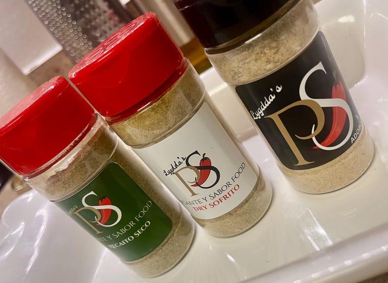 May include: Three spice jars with red lids and labels featuring the text "Lygdda's" and "PS" with a chili pepper graphic. The jars contain a light brown spice blend, with labels that read "Recaito Seco", "Dry Sofrito", and "Adobo".