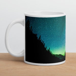 Puede incluir: Taza de cerámica blanca con una silueta negra de un bosque sobre un fondo de cielo estrellado azul y verde.