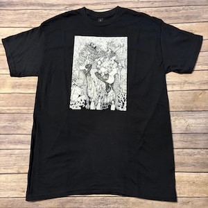 Pode incluir: T-shirt preto com uma impressão gráfica detalhada em preto e branco. O gráfico apresenta várias figuras em um estilo artístico detalhado. A camisa é feita de um material macio e tem um decote redondo clássico.