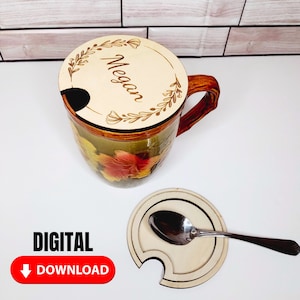 May include: A personalized wooden mug lid with the name "Megan" engraved in a cursive font. The lid has a leaf design around the name. The mug lid is on top of a brown and green mug with a floral design. A wooden coaster with a spoon rest is also shown.