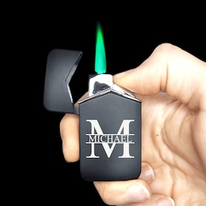 May include: Black lighter with a silver trim and a green flame. The lighter has a personalized inscription that reads "MICHAEL" with a large "M" above the name.