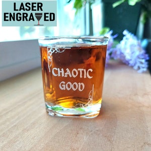May include: A clear glass shot glass with a brown liquid inside. The glass is engraved with the words "Chaotic Good" and a decorative design.