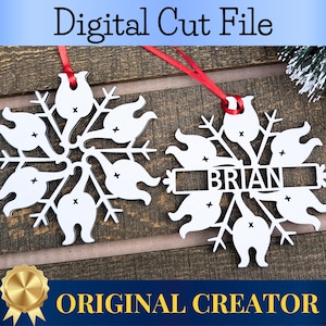 May include: Two white snowflake-shaped ornaments with a personalized name, "BRIAN", on one of them. The ornaments are made of wood and have a cutout design. The ornaments are hanging from red ribbon on a wooden background.