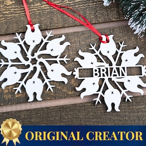 May include: Two white snowflake ornaments with red ribbon hangers. One ornament has the name "BRIAN" cut out. The ornaments are on a wooden surface, with a sprig of evergreen in the background. The ornaments are a festive holiday decoration.