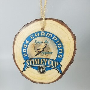 May include: A wooden round ornament with a blue and white design. The design features the text "2004 Champions Tampa Bay Lightning Stanley Cup" with the NHL logo.