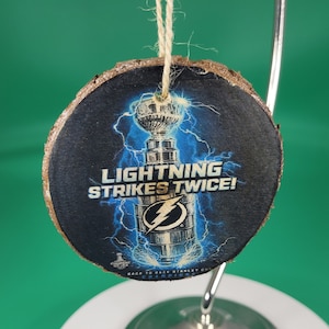 May include: A round wooden ornament with a blue and white image of a hockey trophy and lightning bolts. The text "Lightning Strikes Twice!" is printed on the ornament.