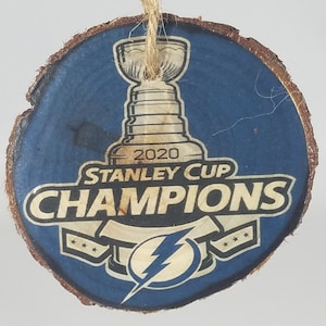 May include: A round wooden ornament with a blue background and a gold Stanley Cup Champions logo. The logo features the Stanley Cup trophy, the Tampa Bay Lightning logo, and the text "2020 Stanley Cup Champions".