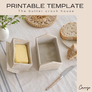 Peut inclure: Un beurrier en céramique en forme de petite maison avec un toit en pente, doté d'une finition beige clair. L'ensemble comprend une base et un couvercle, avec un bloc de beurre jaune à l'intérieur. Un couteau argenté à manche en bois est posé.
