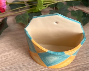 May include: A semi-circular ceramic wall planter featuring a hand-painted diamond pattern in alternating shades of light blue and soft yellow. The interior is a solid cream color, and the back panel includes a single hole for mounting on a wall.