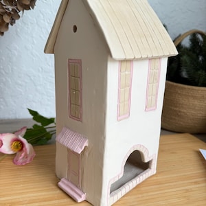 以下が含まれることがあります： オフホワイトとピンクのアクセントが施された、手作りのセラミックハウスの置物。家は傾斜した屋根、窓、小さな日よけのあるドア、装飾的なアーチ型の開口部が特徴です。木製の表面に配置されています。