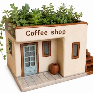 Op de afbeelding: Een keramische plantenbak in de vorm van een koffieshop, met de tekst "Coffee shop" boven de deur. De plantenbak heeft een lichtbeige buitenkant, bruine randen en een blauwe deur. Hij is gevuld met diverse groene planten en vetplanten.