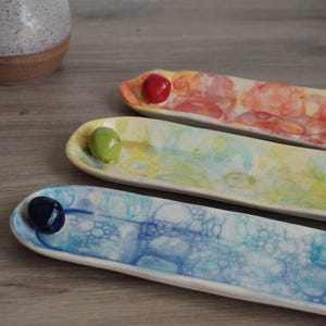 May include: Three handmade ceramic serving dishes. Each dish is long and narrow, with a rounded end and a small, colorful bead. The dishes are painted with watercolor-like patterns in shades of red, yellow, and blue.