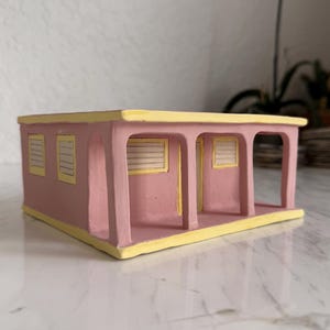 Könnte beinhalten: Ein rosa-gelbes Keramik-Miniaturgebäude mit Veranda. Das Gebäude hat drei Fenster mit horizontalen Jalousien und ein gelbes Dach. Die Veranda wird von vier gelben Säulen getragen. Das Gebäude steht auf einer weißen Marmoroberfläche.