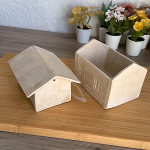 Puede incluir: Dos recipientes de cerámica beige con forma de casa. Uno es una casa cerrada con tejado y la palabra "Cute" escrita en el frente. El otro es un recipiente abierto con detalles de ventana. Ambos están sobre una superficie de madera.
