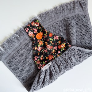 May include: A grey kitchen towel with a black and orange floral print hanging loop. The loop has a button and the text "Tinka" is visible on the loop.