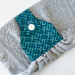 May include: A grey kitchen towel with a teal and yellow patterned fabric loop attached. The loop has a white button and the towel has a white tag that says "Tinka".
