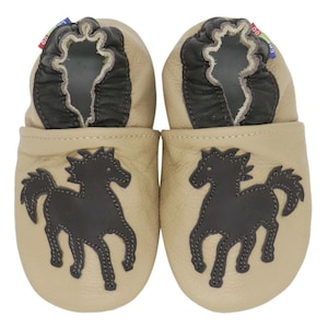 May include: A pair of beige leather baby shoes with black horse appliqués. The shoes have a black leather trim around the opening and a soft, flexible sole. Small red and blue tags are attached to the shoes.