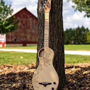 May include: A wooden acoustic guitar with a paisley design burned into the body. The guitar is leaning against a tree trunk in a grassy field.