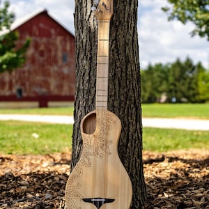 May include: A wooden acoustic guitar with a paisley design burned into the body. The guitar is leaning against a tree trunk in a grassy area.