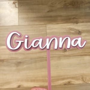 May include: A pink acrylic cake topper with the name "Gianna" written in white lettering.