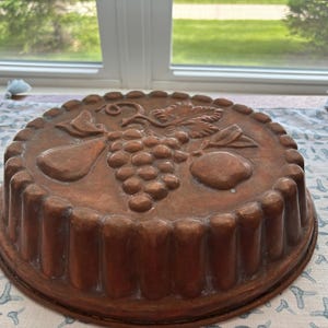 May include: A round copper-coloured cake mould featuring a moulded relief design on its top surface. The design depicts a cluster of grapes, a pear, and an apple with leaves and vines. The mould has a fluted, scalloped edge around its perimeter.