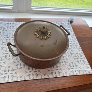 May include: A round copper pot with a matching lid, featuring two twisted metal side handles and a decorative brass-toned finial on top. The pot has a warm, metallic finish with visible signs of age and use, showcasing a classic, functional design.
