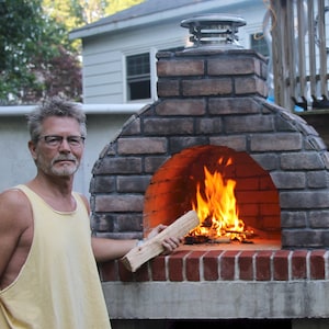 May include: A brick pizza oven with a fire burning inside. The oven is constructed from red and grey bricks and features a stainless steel chimney. A person is holding a piece of wood, ready to add it to the flames.