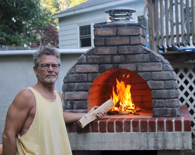 May include: A brick pizza oven with a fire burning inside. The oven is constructed from red and grey bricks and features a stainless steel chimney. A person is holding a piece of wood, ready to add it to the flames.