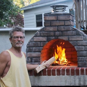 Puede incluir: Un hombre se encuentra frente a un horno de pizza exterior de ladrillo con fuego ardiendo en su interior. Sostiene un trozo de le&ntilde;a en su mano derecha.
