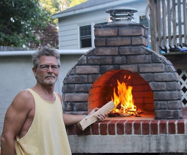 Puede incluir: Un hombre se encuentra frente a un horno de pizza exterior de ladrillo con fuego ardiendo en su interior. Sostiene un trozo de le&ntilde;a en su mano derecha.