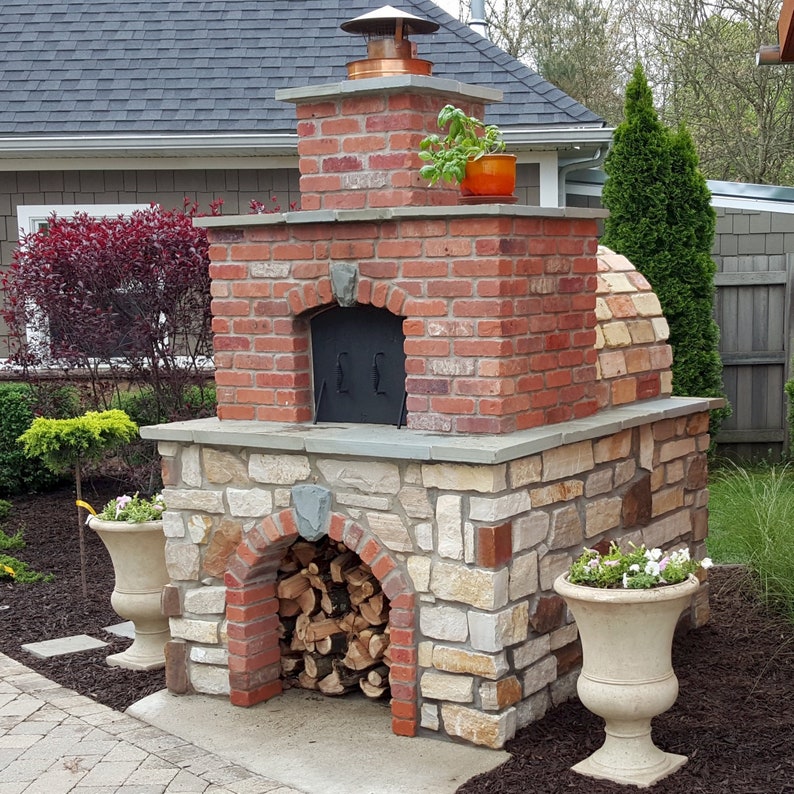 May include: An outdoor pizza oven constructed from red brick and stone, featuring a copper chimney cap. The oven has a brick chimney and a stone base. A small orange pot with basil sits on top. Firewood is stacked in the lower opening. Two decorative urns with flowers.