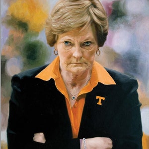 May include: A woman with short blonde hair wearing a black blazer over an orange collared shirt with a 'T' on the right side. She has her arms crossed and is looking directly at the viewer.