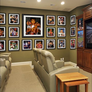 May include: A home theater room with a wall of framed sports and boxing prints, two beige reclining chairs, and two end tables. The room has a brown carpet and a brown entertainment center with a television.