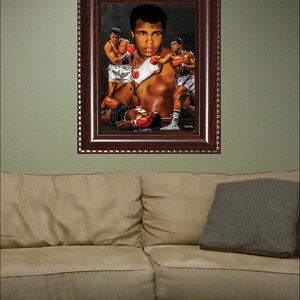 May include: A framed painting of a boxing match featuring Muhammad Ali. The painting depicts Ali in a dynamic pose, throwing a punch, with two other boxers in the background. The painting is set against a dark background and is framed in a dark wood frame.