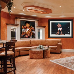 May include: A living room interior with a brown leather sectional sofa, a glass-top coffee table, and two framed boxing prints featuring Muhammad Ali. The prints are hung on a wall painted a warm orange color. The room has hardwood floors and a rug with a floral pattern.